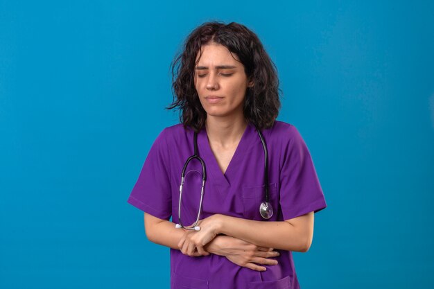 Young woman nurse in medical uniform and with stethoscope looking unwell touching stomach having pain standing