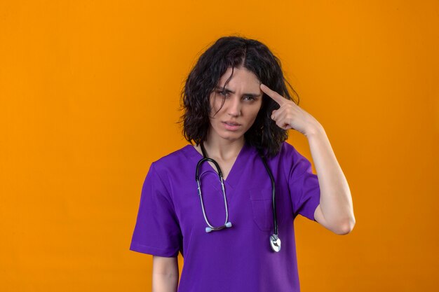 Young woman nurse in medical uniform and with stethoscope pointing temple with finger concentrating hard on an idea with a serious look