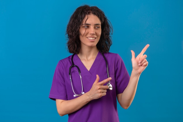 Young woman nurse in medical uniform and with stethoscope smiling cheerfully looking aside pointing with fingers to the side standing