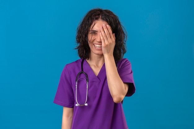 医療制服と聴診器で笑顔と驚きの立っている目を覆っている顔に手で笑う若い女性看護師