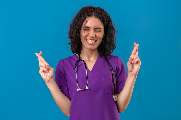 Young woman nurse in medical uniform and with stethoscope standing with closed eyes raises fingers crossed makes desirable wish