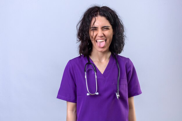 Young woman nurse in medical uniform and with stethoscope sticking tongue out happy with funny expression standing