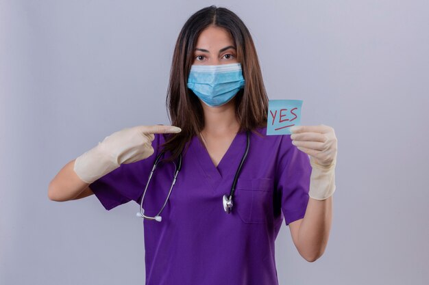 Young woman nurse wearing medical uniform protective mask gloves and with stethoscope holding reminder paper with yes word pointing to it with index finger