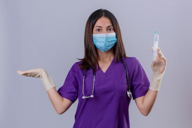 Young woman nurse wearing medical uniform protective mask gloves and with stethoscope holding syringe looking clueless and confused standing with arm raised