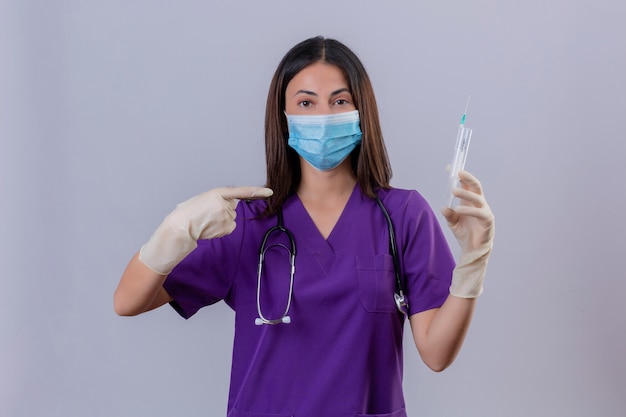 Young woman nurse wearing medical uniform protective mask gloves and with stethoscope holding syringe pointing to it with index finger
