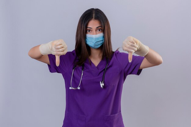 Young woman nurse wearing medical uniform protective mask gloves and with stethoscope showing thumbs down