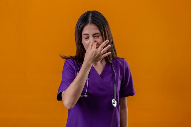 Young woman nurse wearing uniform and stethoscope closing nose with fingers feels unpleasant scent suffers from stench