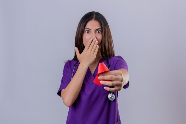 Young woman nurse wearing uniform and with stethoscope holding red ribbon a symbol of the fight against aids shocked covering mouth with hand