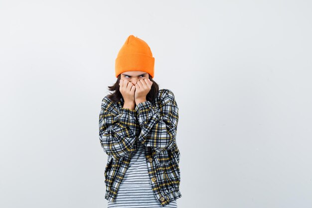 Молодая женщина в оранжевой шляпе клетчатой рубашке держит кулаки возле лица, выглядя испуганной