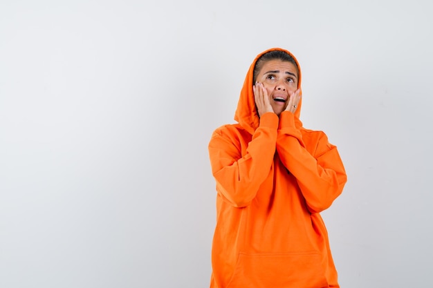 Young woman in orange hoodie holding hands near mouth as calling someone and looking focused 