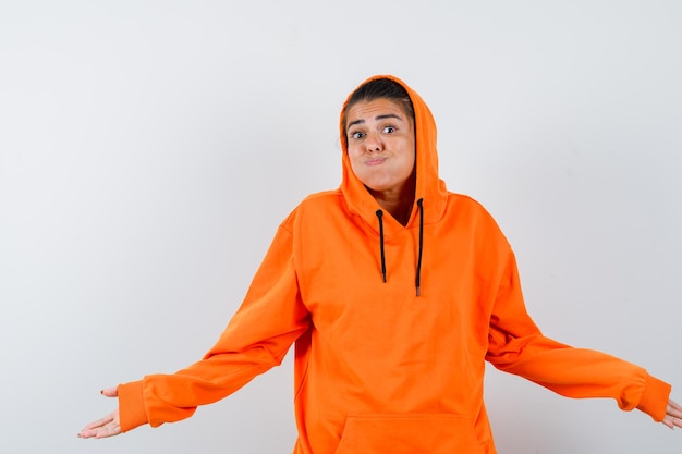 Young woman in orange hoodie stretching hands in questioning manner and looking baffled 