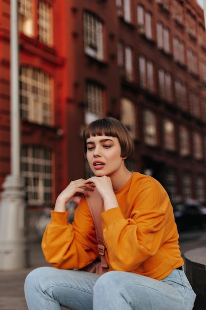 Young woman in orange sweatshirt and jeans sits outdoors Shorthaired girl in bright sweater and denim pants poses outside