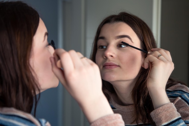 A young woman paints her eyelashes with mascara in front of a mirror