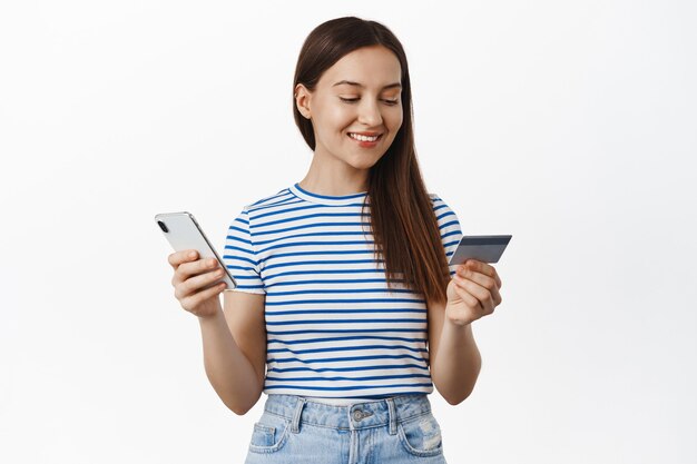 신용 카드와 휴대 전화로 지불하는 젊은 여성, 웃고 편안한 표정, 인터넷 상점에서 smth를 구입, 스마트폰 응용 프로그램에서 구입, 흰 벽 위에 서 있습니다.