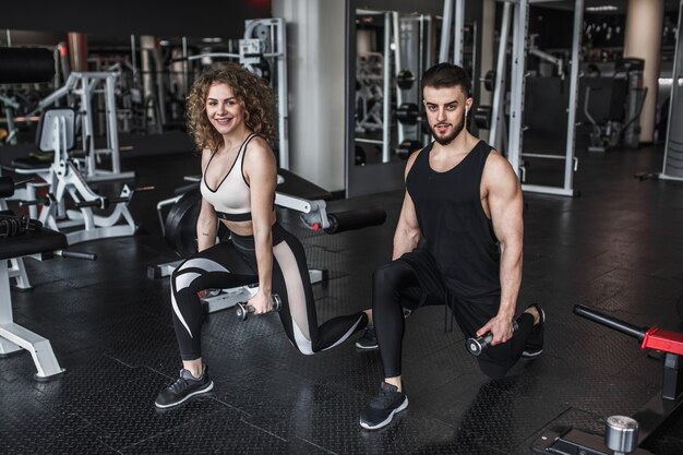 Young woman and personal trainer with dumbbell squats in gym