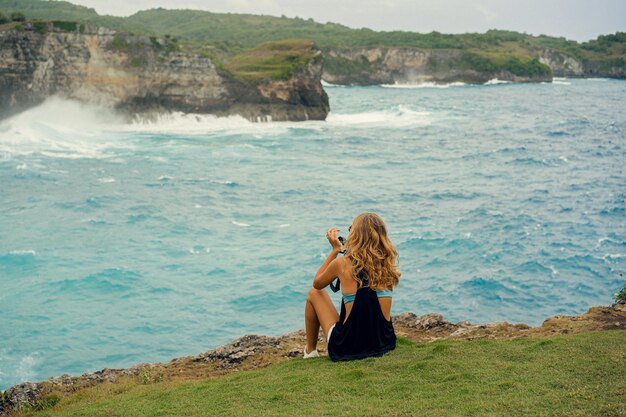 Young woman photographer traveler with a camera on the edge of a cliff takes pictures of nature