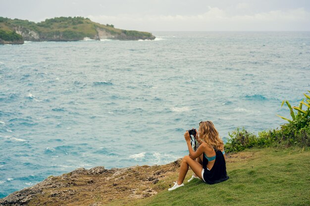 Young woman photographer traveler with a camera on the edge of a cliff takes pictures of nature