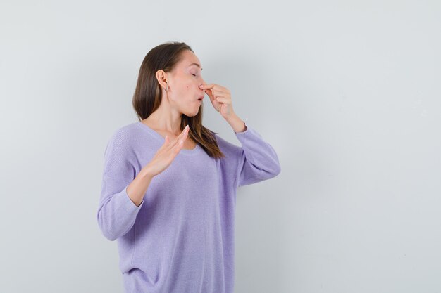 Young woman pinching her nose in lilac blouse and looking disgusted 