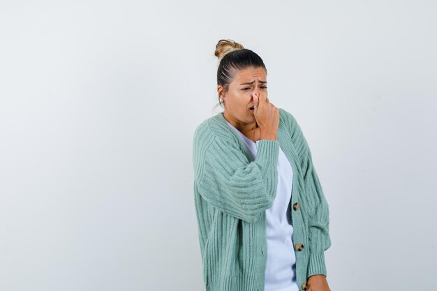 Young woman pinching nose due to bad smell in white shirt and mint green cardigan and looking harried