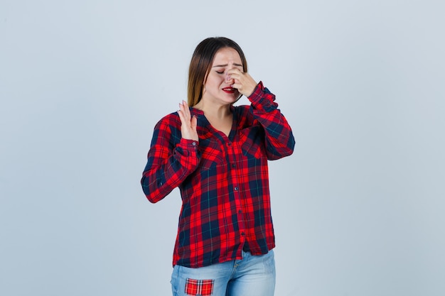 Young woman pinching nose, showing stop sign while crying in checked shirt, jeans and looking morose. front view.