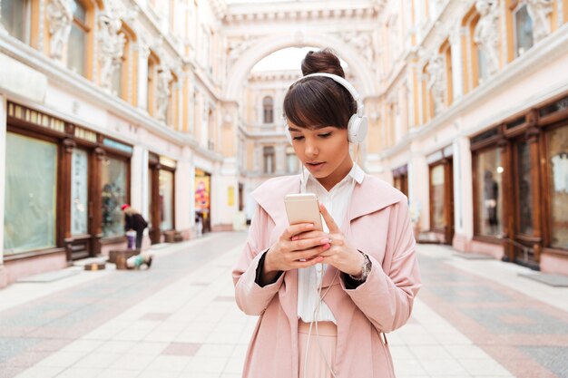 Young woman in pink coat listening to music with headphones outside