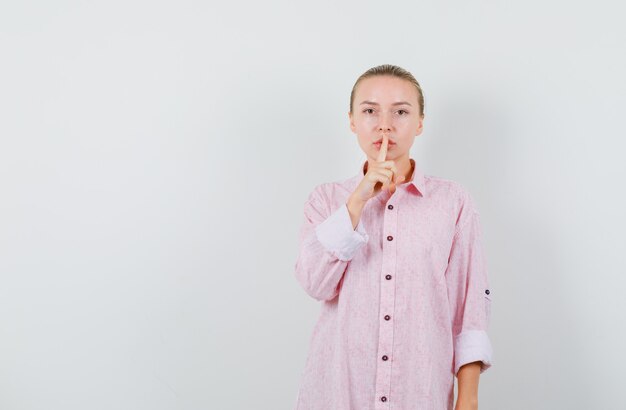 沈黙のジェスチャーを示し、注意深く見ているピンクのシャツの若い女性