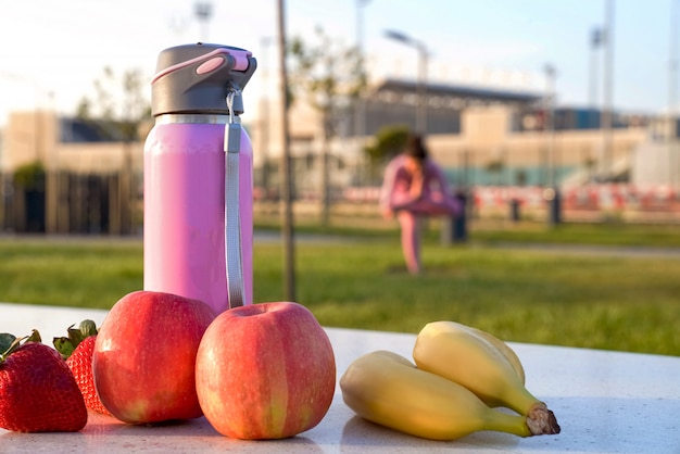 young woman in pink shirt and trousers on the grass inside park fruits bottle meditating and doing yoga in different poses