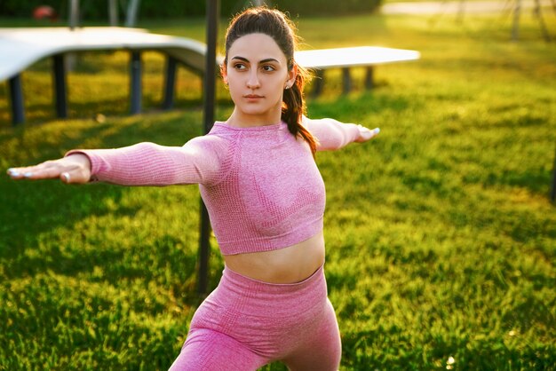 Foto gratuita giovane donna in camicia rosa e pantaloni sull'erba all'interno del parco meditando e facendo yoga in diverse pose