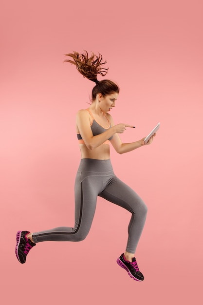 Free photo young woman over pink studio wall using tablet gadget while jumping