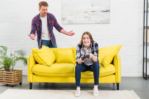 黄色のソファをすくめて後ろに立っている彼女のボーイフレンドと一緒にビデオゲームをプレイ若い女性