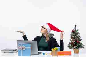 Foto gratuita giovane donna che gioca con il cappello di babbo natale e gli occhiali seduti a un tavolo con un albero di natale e un regalo