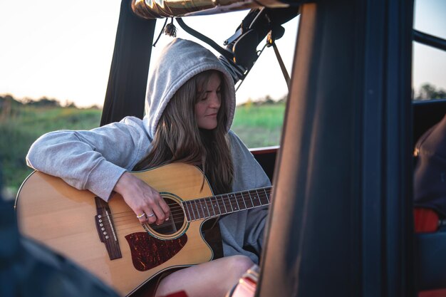 한 젊은 여성이 자연 속에서 차 트렁크에서 어쿠스틱 기타를 연주한다