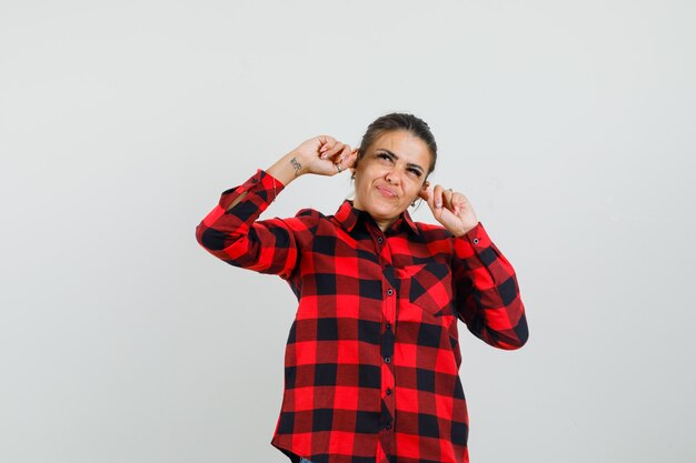 Young woman plugging ears with fingers in checked shirt and looking annoyed. front view.