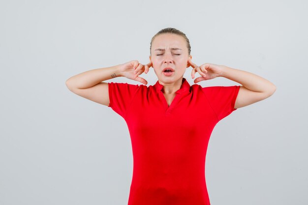 Молодая женщина затыкает уши пальцами в красной футболке и выглядит раздраженной