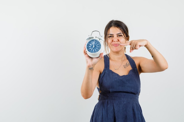 Young woman pointing at alarm clock in dress and looking punctual