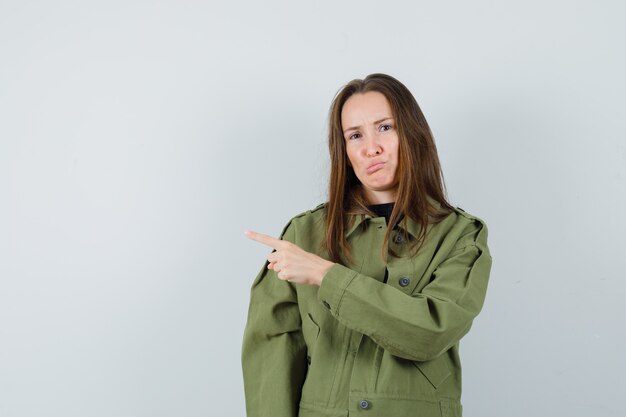 Young woman pointing aside in green jacket and looking unhappy , front view.