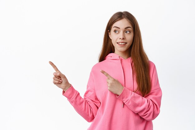 Young woman pointing aside, looking at left copy space with curious expression, checking out promo, standing against white wall