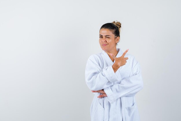 Young woman pointing aside while blinking in bathrobe and looking confident