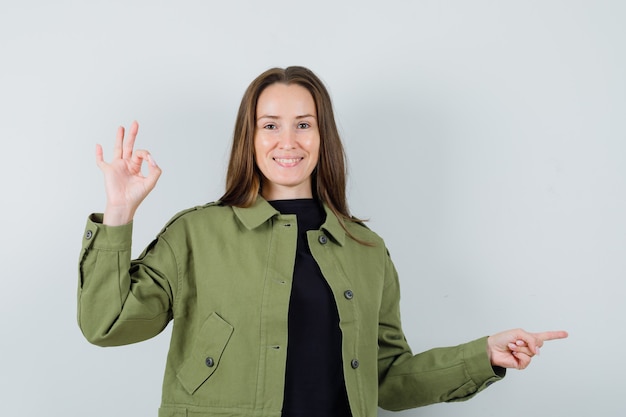 Young woman pointing aside while showing ok gesture in green jacket and looking glad. front view.