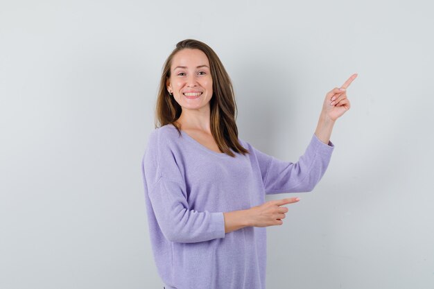 Young woman pointing aside while smiling in lilac blouse and looking cheery. front view.