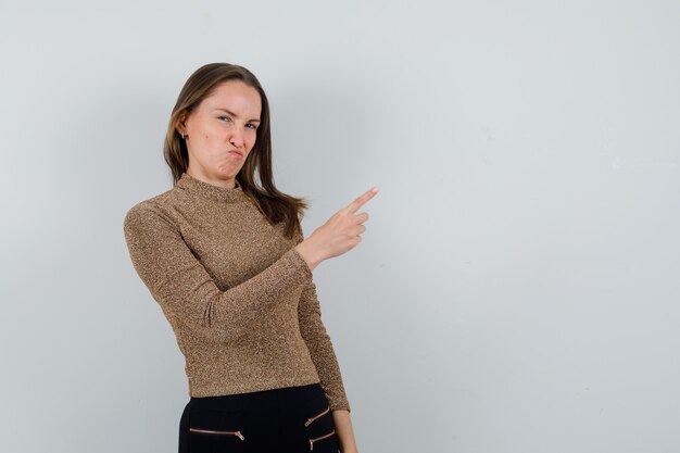 Young woman pointing aside while souring her face in golden blouse