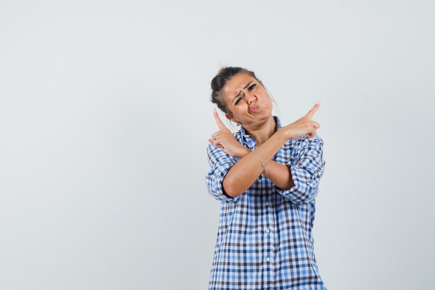 Young woman pointing aside with crossed arms in checkered shirt and looking confused.