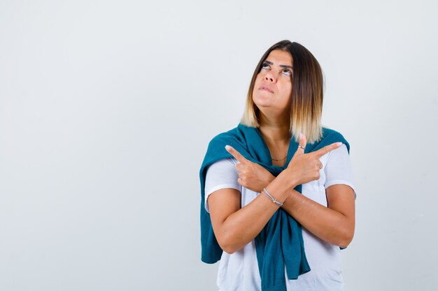 Young woman pointing aside with crossed arms in white t-shirt and looking puzzled , front view.