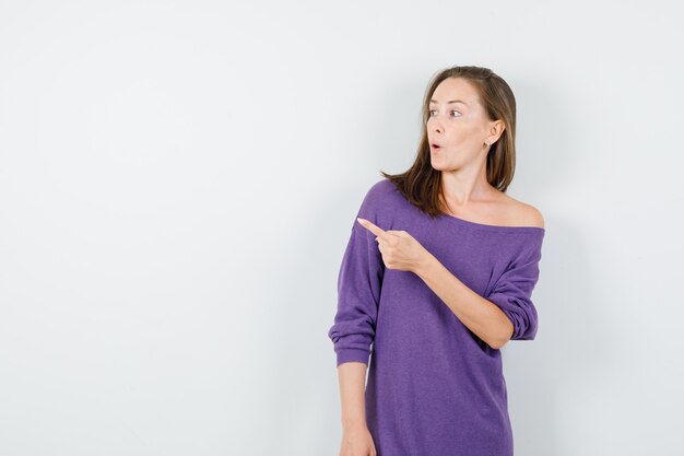 Young woman pointing away in violet shirt and looking amazed. front view.