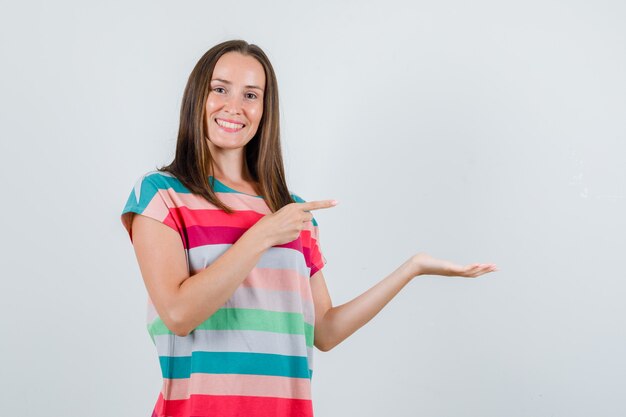 Young woman pointing away with palm spread aside in t-shirt and looking cheery , front view.