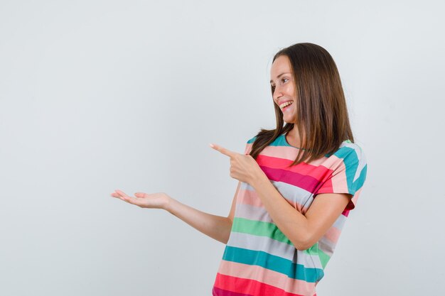 Young woman pointing away with palm spread aside in t-shirt and looking cheery , front view.