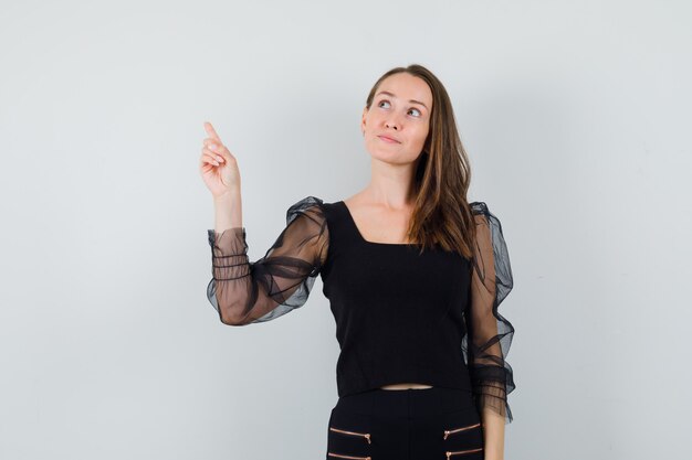 Young woman pointing back in black blouse and looking focused. front view.