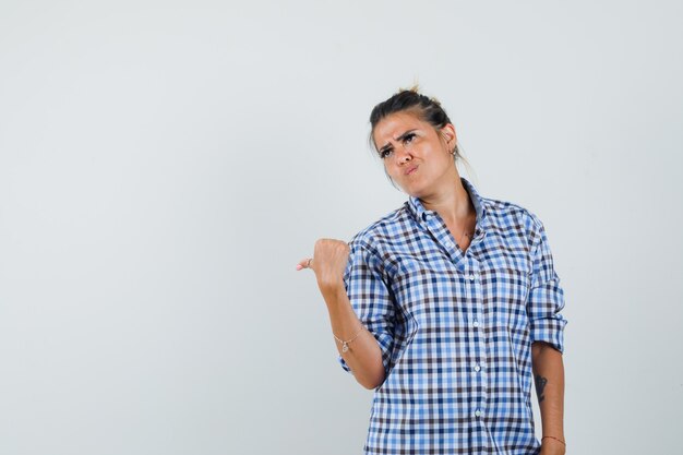 Young woman pointing back in checkered shirt and looking reluctant