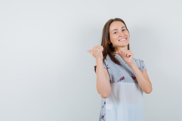 Young woman pointing back in t-shirt and looking happy. front view.