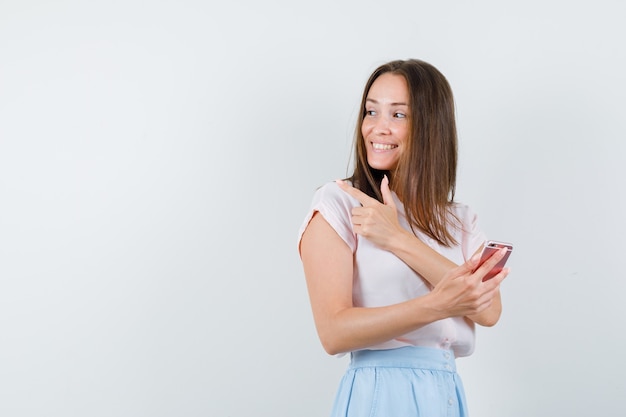 Young woman pointing back while holding mobile phone in t-shirt, skirt and looking cheerful , front view.
